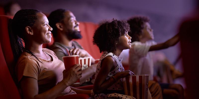 Family sits together smiling in movie theatre eating popcorn