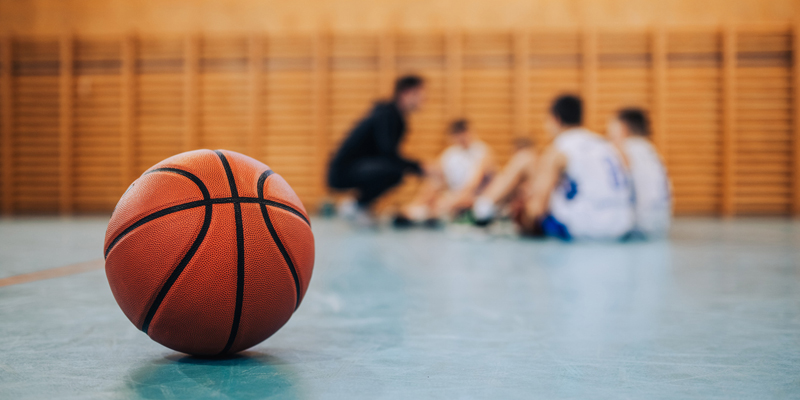 A basketball on the floor of a gym with a group of teen basketball players huddle together in the background